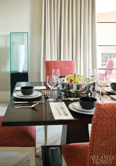 Dark wood dining table set with black bowls, white plates, silverware, wine glasses, and a silver bowl of fruit. Red patterned chairs surround the table.