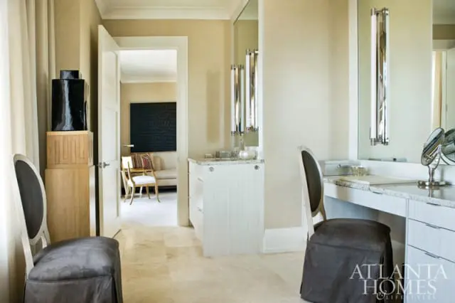 Neutral toned dressing room with textured wallpaper, marble countertops, two upholstered chairs, and a doorway revealing a living area with a black textured artwork.