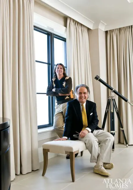 Two men posing in a room with large windows and beige curtains. One man sits on a bench wearing a dark suit jacket, yellow tie, and light pants. The other man stands behind him wearing a sweater and light pants. A telescope on a tripod is visible to the right.