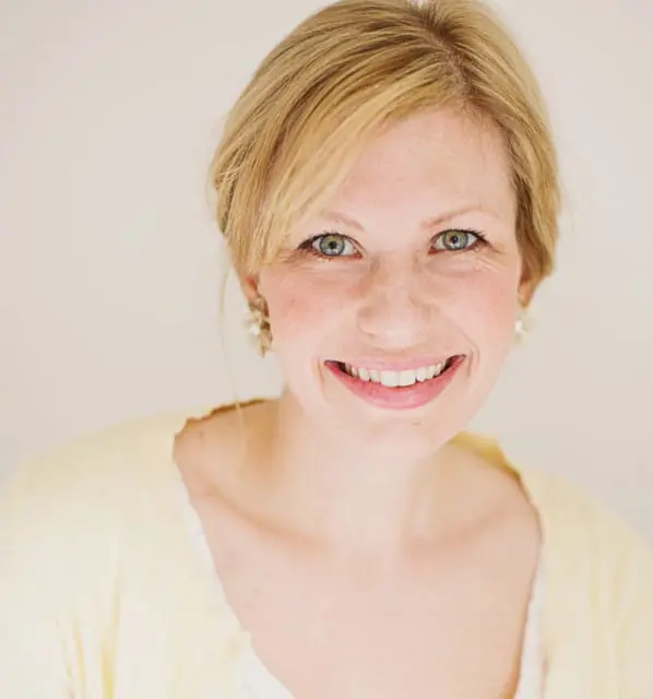 Close-up portrait of a smiling woman with blonde hair, green eyes, and freckles, wearing a light yellow cardigan and pearl earrings.
