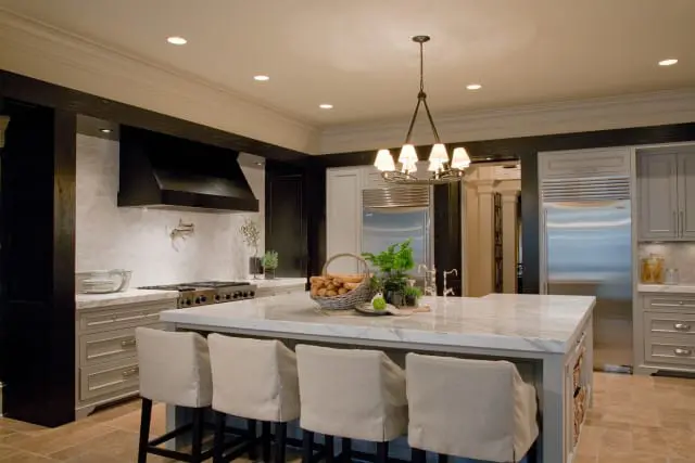 Kitchen island with marble countertop and four upholstered stools, facing a stainless steel range with a black hood and white tile backsplash.