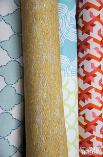 Close-up of four rolled fabrics: one with a blue quatrefoil pattern, one with a textured yellow pattern, one with a blue wave pattern, and one with an orange geometric pattern.