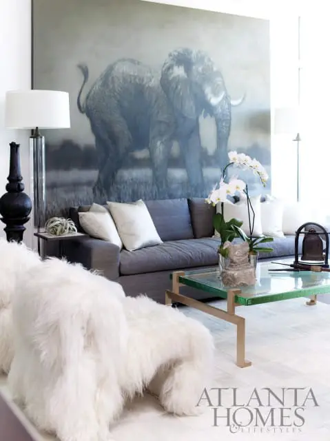 Living room with a large painting of an elephant, gray sofa with white pillows, white furry ottoman, and a glass coffee table with a plant.
