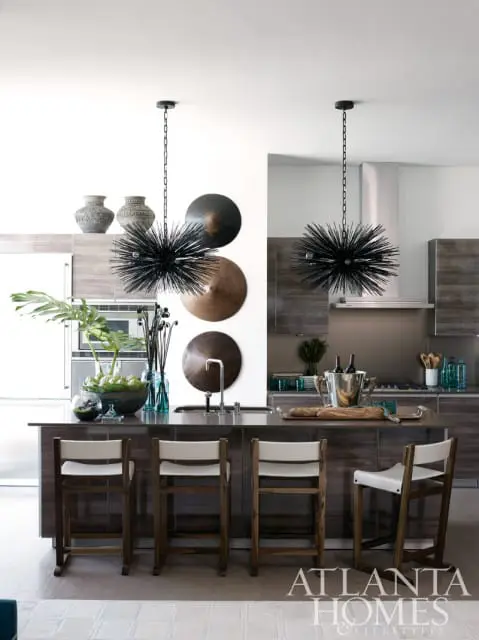 Modern kitchen island with dark wood cabinetry and white leather stools, featuring sputnik chandeliers, decorative vases, and a stainless steel sink.