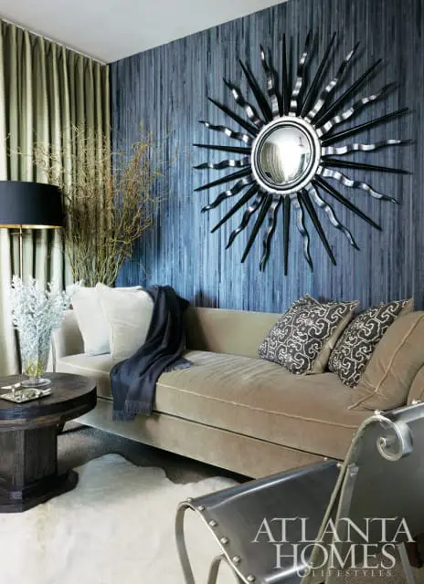 Living room with a tan velvet sofa, blue textured wallpaper, a sunburst mirror, and a dark wood side table with a white rug.
