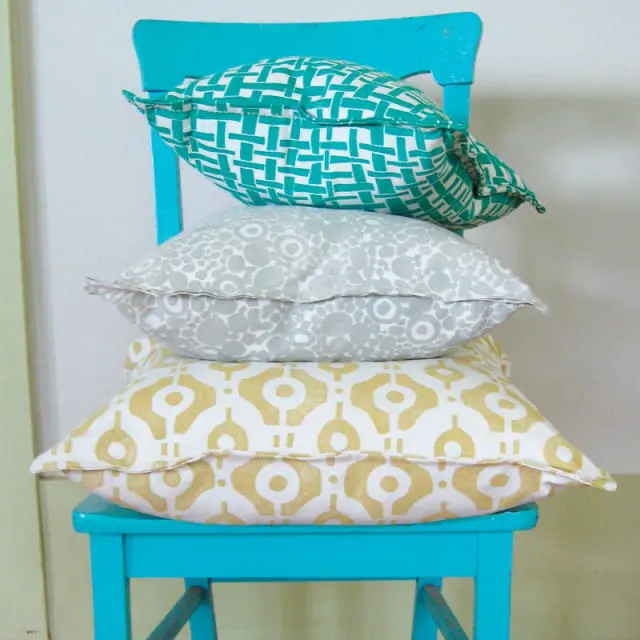 Stack of three decorative pillows on a bright teal chair. Top pillow has a white and teal geometric pattern. Middle pillow has a white and gray abstract floral pattern. Bottom pillow has a white and gold geometric pattern.