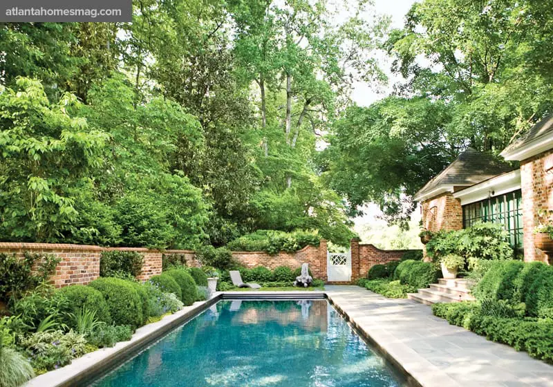 Rectangular swimming pool with turquoise water, surrounded by manicured hedges, brick walls, and lush trees. A stone patio leads to a brick building.