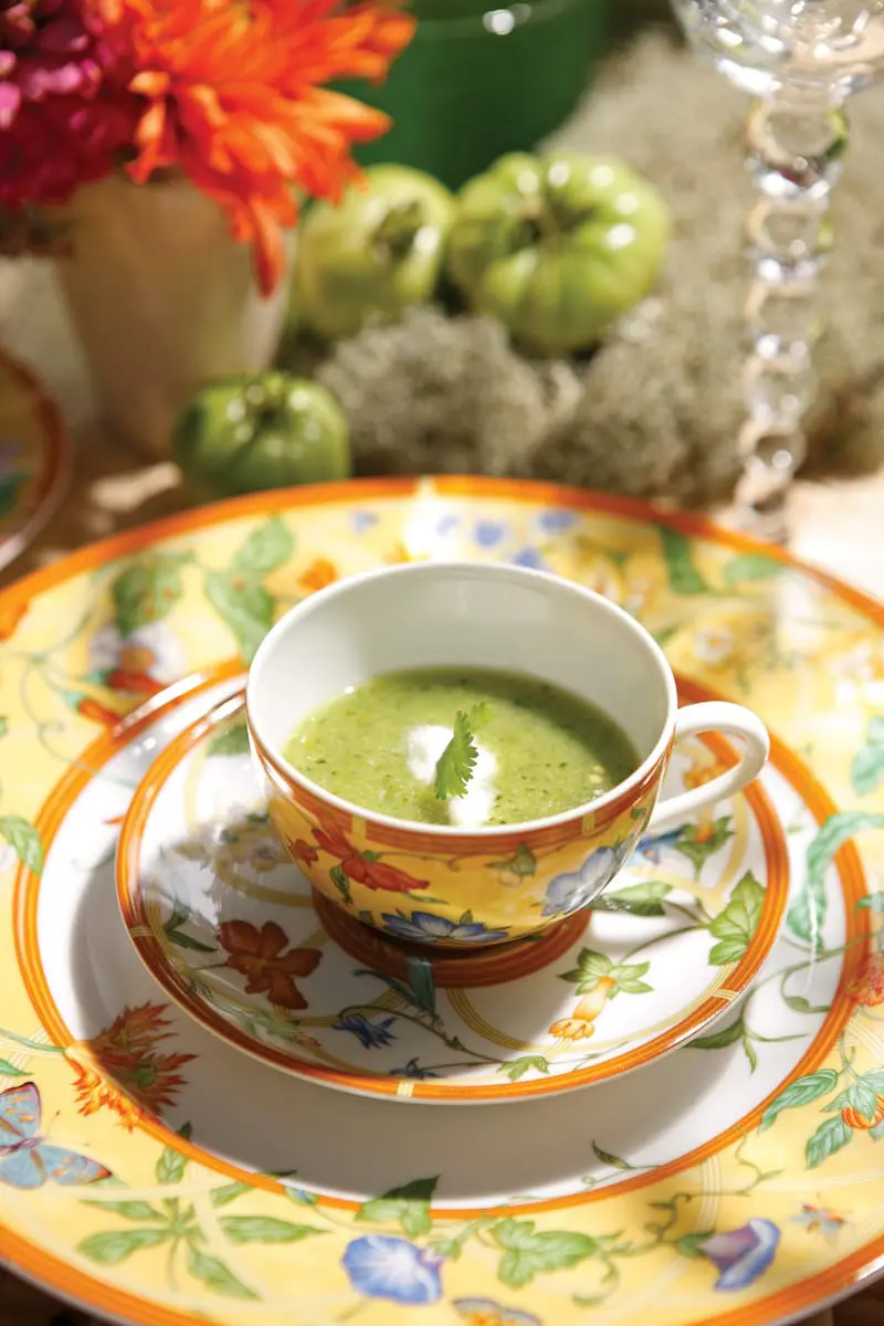 Close-up of a white soup cup filled with green soup, garnished with a swirl of cream and a cilantro sprig, resting on a floral patterned plate.