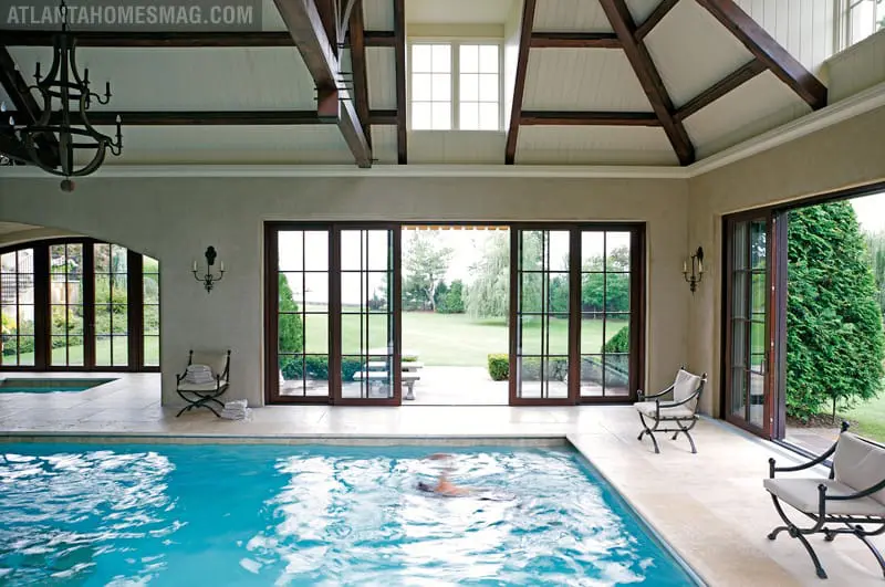 Indoor pool with bright blue water, surrounded by light stone tile and large glass doors opening to a green lawn. Exposed wooden beams support the vaulted ceiling.