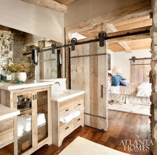 Rustic bathroom with a reclaimed wood vanity, stone accent wall, and a sliding barn door leading to a bedroom with a king-sized bed and blue pillows.