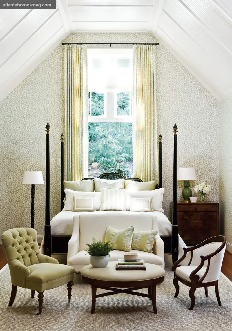Bedroom with a four-poster bed, tufted armchair, and upholstered bench. Walls are covered in a subtle floral wallpaper, and the vaulted ceiling features white wood paneling.