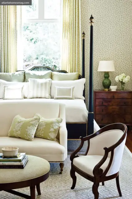 Bedroom with a four-poster bed, cream sofa, armchair, and a dark wood dresser with a green lamp. Walls are covered in a light floral wallpaper.
