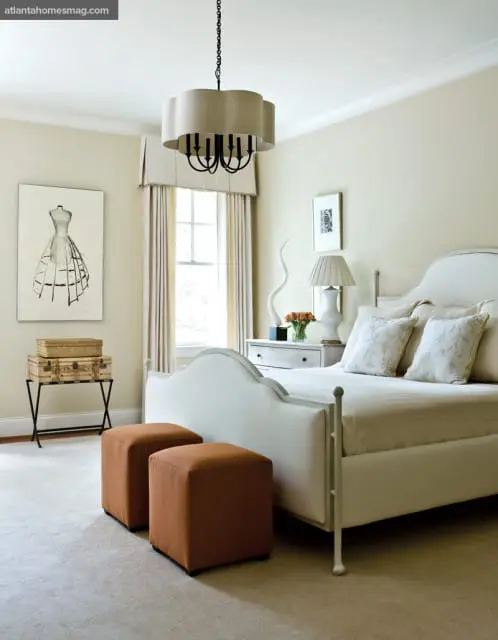 Cream bedroom with a white upholstered bed, two rust-colored ottomans, a dresser with a lamp and floral arrangement, and artwork of a dress form on the wall.