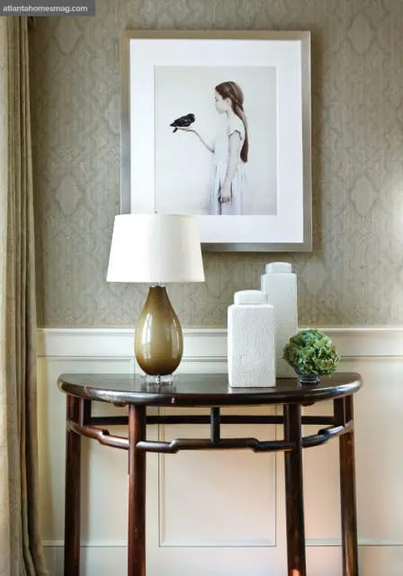 Console table with a brown glass lamp, two white textured vases, and a small green plant under a framed artwork of a girl holding a bird.