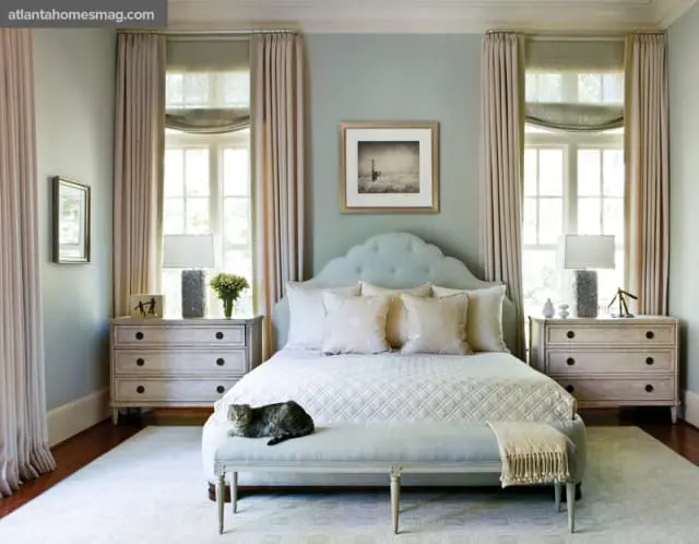 Light blue tufted headboard bed with white quilted bedding, flanked by two light wood dressers and tall beige curtains. A tabby cat rests on a light blue bench at the foot of the bed.