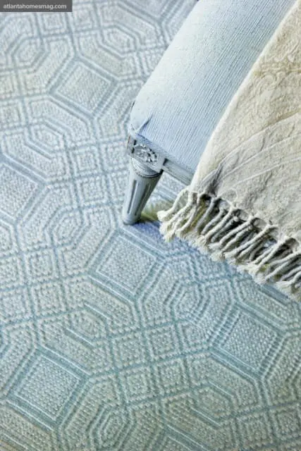 Close-up of a light blue upholstered chair leg with carved details, resting on a light blue geometric patterned rug. A cream-colored throw with fringe is draped over the chair.