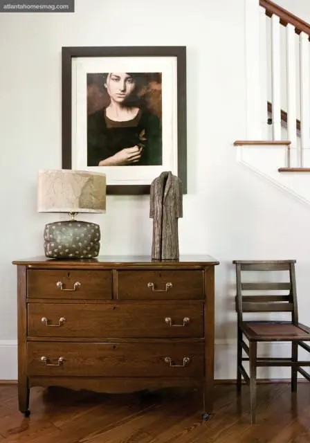Dark wood dresser with brass hardware, topped with a patterned lamp and a wood sculpture. A framed portrait hangs above. A wooden chair sits to the right.