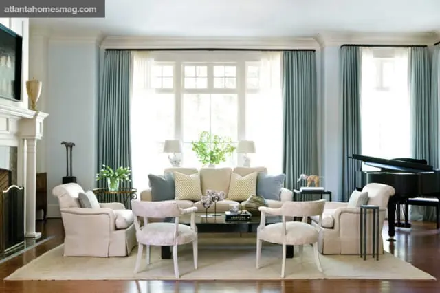 Living room with a cream sofa, upholstered armchairs, and accent chairs arranged around a dark coffee table. Light blue curtains frame a large window, and a black grand piano is visible on the right.