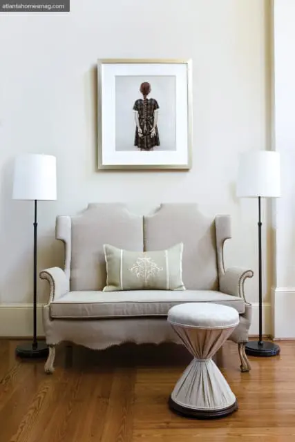 Neutral toned loveseat with decorative pillow, flanked by two floor lamps, in front of framed artwork of a girl with braided hair.