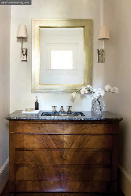Powder room with a dark marble countertop vanity featuring a wooden dresser base, brass fixtures, and a large framed mirror. White orchids in a glass vase sit on the counter.
