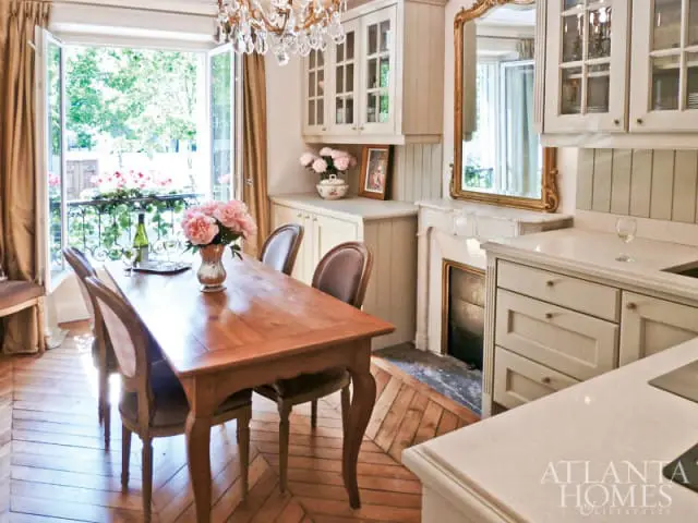 Cream kitchen with a wooden dining table, upholstered chairs, a fireplace with a gold mirror, and a balcony view with pink flowers.