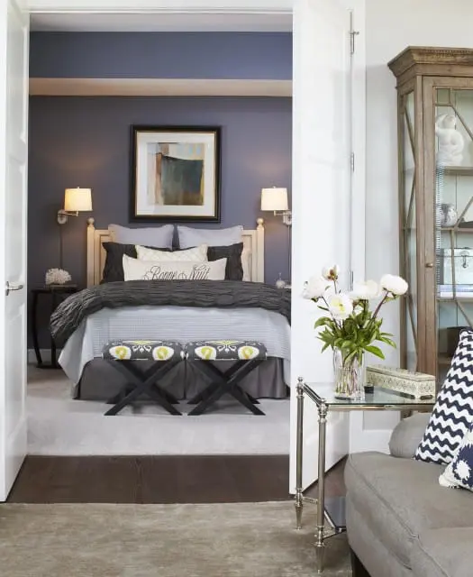 Bedroom with dark blue walls, a king-sized bed with gray bedding, two wall-mounted lamps, and two patterned ottomans. A glass-top table with a vase of white peonies sits in the foreground.