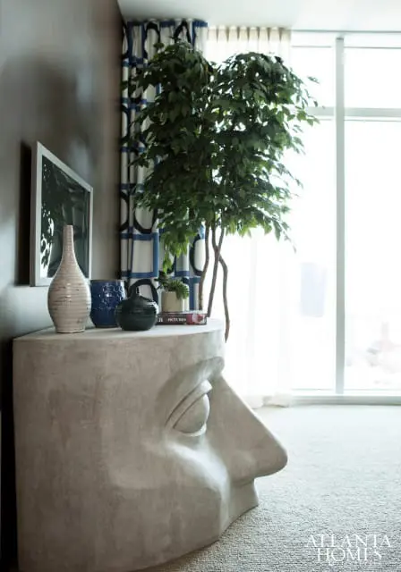 Interior scene with a console table shaped like a human head profile, topped with vases and a plant. Blue and white patterned curtains hang behind a large potted tree.