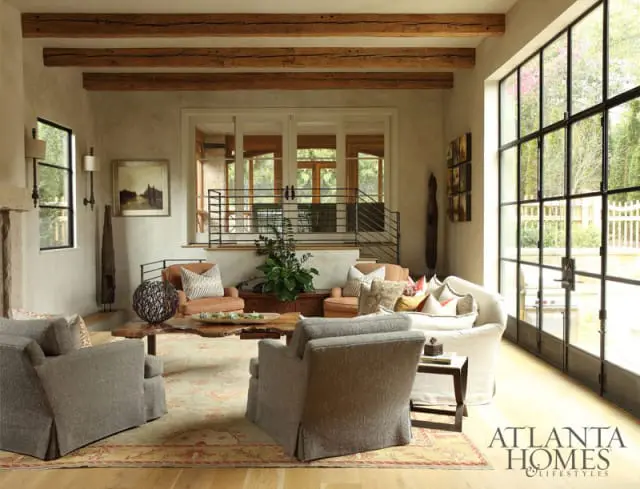 Living room with exposed wooden beams, large steel-framed windows, neutral-toned slipcovered furniture, a live-edge wood coffee table, and a patterned area rug.