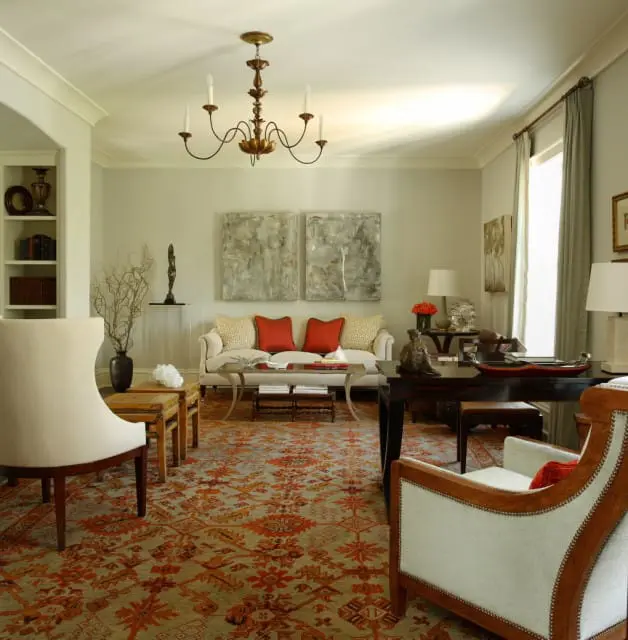 Living room with a cream sofa, two red accent pillows, and a patterned rug in shades of orange and grey. A dark wood desk and chairs are also visible.