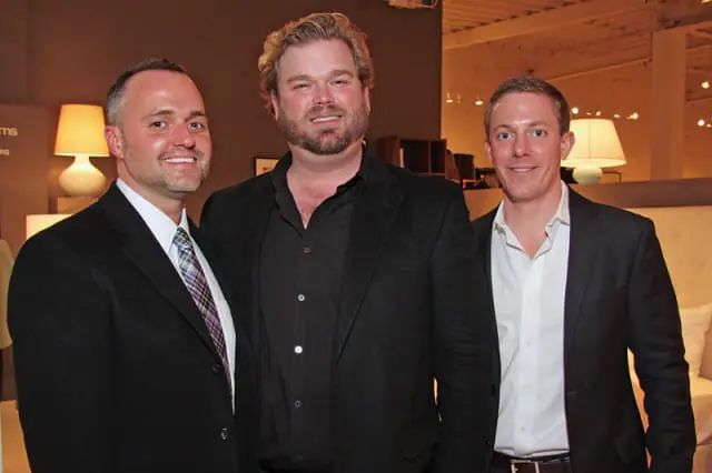 Three men in suits smile at the camera. The man on the left wears a black suit with a patterned tie. The man in the center wears a black suit jacket over a black open-collar shirt. The man on the right wears a dark suit jacket over a white shirt.
