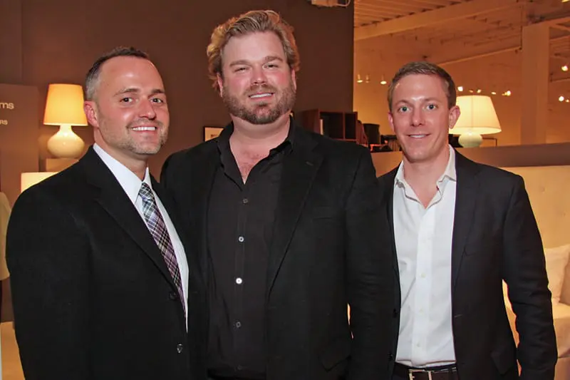 Three men in suits smile at the camera. The man on the left wears a black suit with a patterned tie. The man in the center wears a black suit jacket over a black open-collar shirt. The man on the right wears a dark suit jacket over a white shirt.