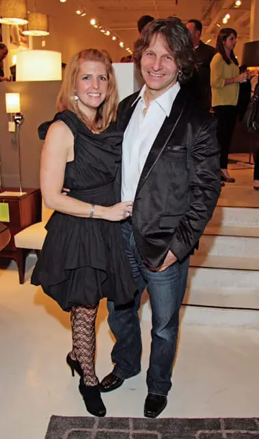 A smiling woman in a black ruffled dress and fishnet stockings stands next to a smiling man in a black satin-finish blazer and jeans.