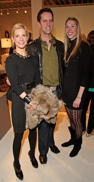 A woman in a dark gray dress and large silver necklace stands with a man in a green shirt and leather jacket, and another woman in a black dress and patterned tights.