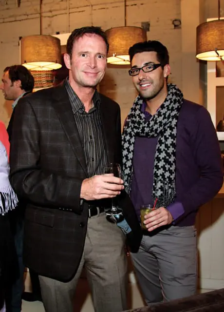 Two men standing together, one in a plaid blazer and striped shirt, the other in a purple sweater and patterned scarf, both holding drinks.