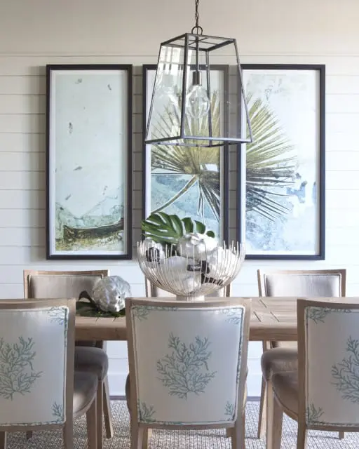 Dining room with light wood table and chairs upholstered in light fabric with blue coral pattern. Large framed botanical prints hang on a shiplap wall above the table.