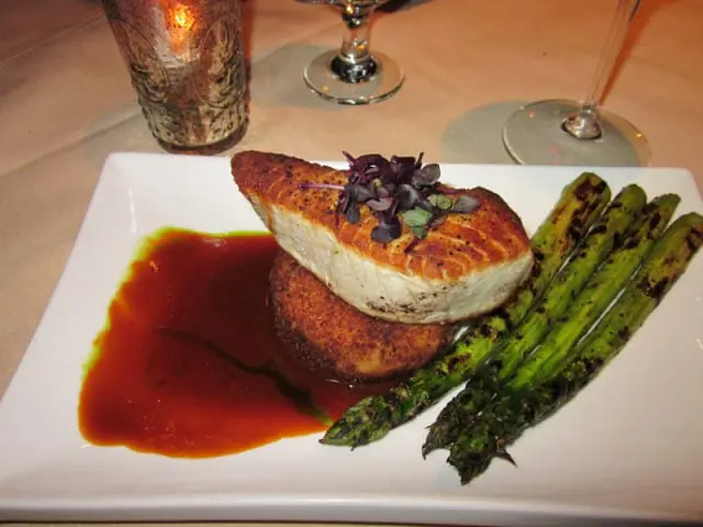 Seared fish fillet served with a round breaded patty, red sauce, and grilled asparagus on a white rectangular plate.