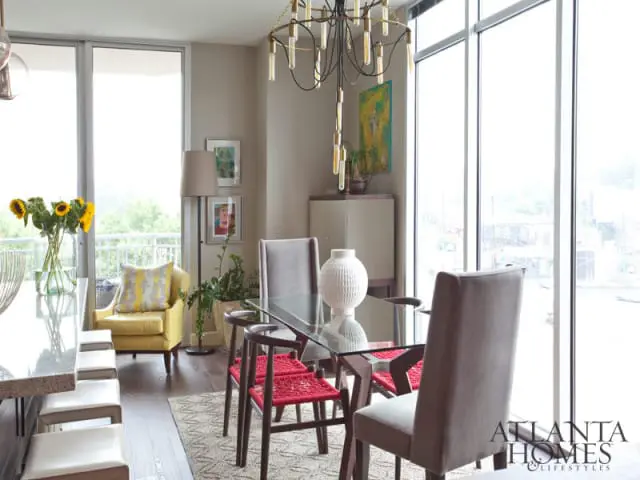Dining area with glass-top table, upholstered chairs, red woven chairs, yellow armchair, sunflowers, and large windows overlooking a city view.