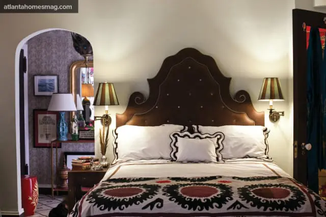 Bedroom with a large brown upholstered headboard, white bedding with decorative trim, and a patterned throw blanket. Striped lampshades flank the bed.