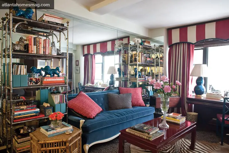 Blue velvet sofa with patterned red and gray pillows in a living room with mirrored walls, a chrome etagere filled with books and decor, and striped curtains.