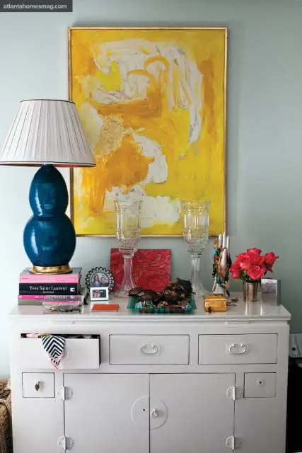 White dresser topped with a blue lamp, books, abstract yellow art, crystal vases, jewelry, and a bouquet of pink roses.