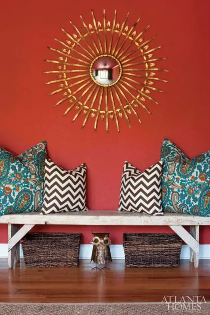 Red accent wall with a gold sunburst mirror above a rustic wooden bench adorned with paisley and chevron patterned pillows, flanked by woven baskets.