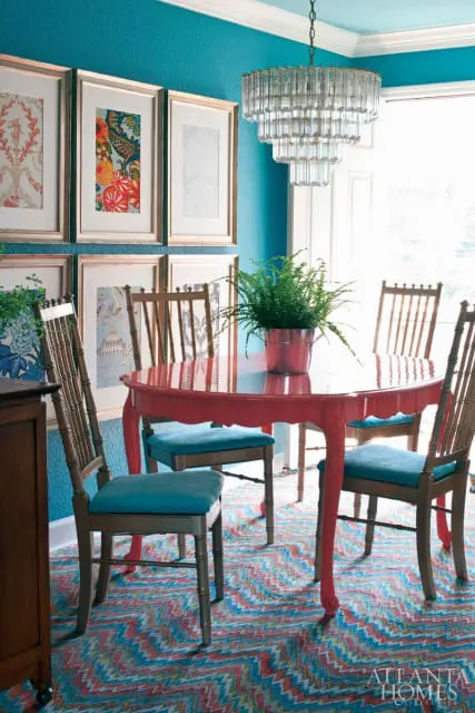Dining room with teal walls, a coral pedestal table with teal upholstered chairs, and a colorful chevron rug. Framed artwork hangs on the wall above a dark wood cabinet.
