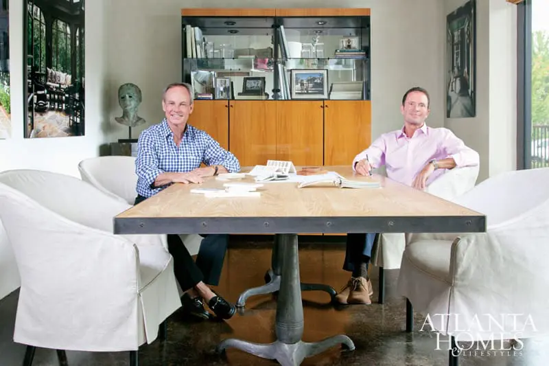 Two men sit at a wooden table in a room with a large wooden cabinet filled with books and decorative items. One man wears a blue gingham shirt, the other a pink dress shirt.