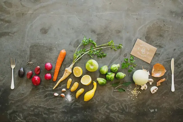 A flat lay of fresh ingredients including parsnips, carrots, potatoes, lemons, Brussels sprouts, onion, mushrooms, and herbs arranged on a dark textured surface with a fork and knife.