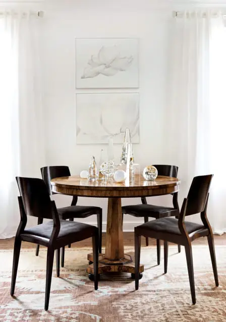 Dining room with a round wooden table set with glass decorative objects and four dark upholstered chairs. Two framed floral art pieces hang on the wall above the table.