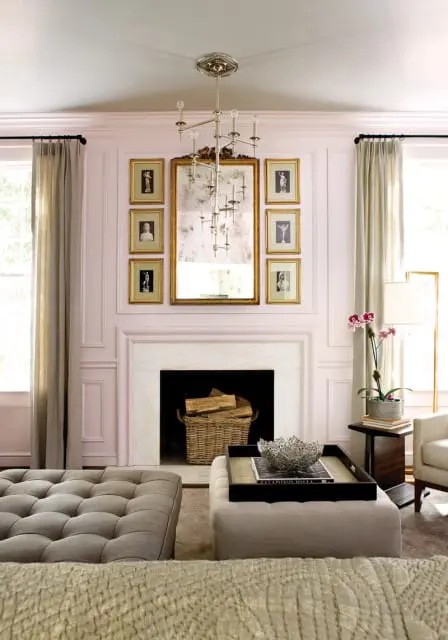 Living room with a pale pink paneled wall, marble fireplace with a basket of logs, a large antique mirror surrounded by framed art, and tufted gray and beige ottomans.