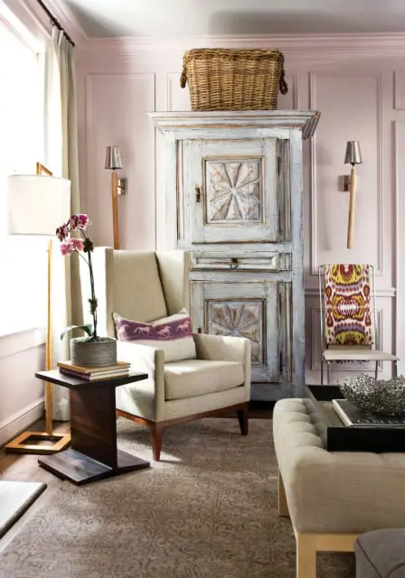 Living room with a light beige wingback chair, a distressed blue armoire, and a patterned rug. A floor lamp and wall sconces provide lighting.