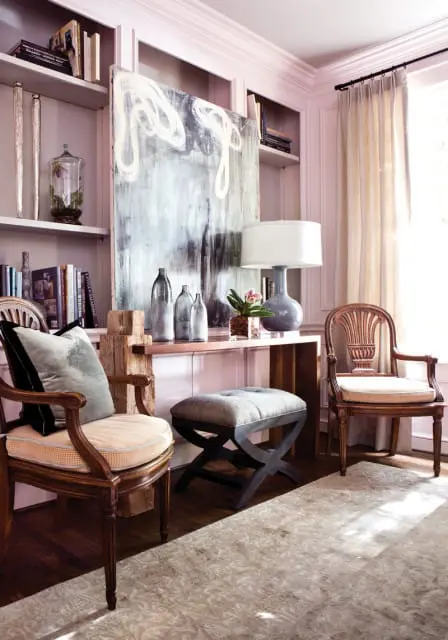 Living room with built-in bookshelves painted pale pink, featuring two wooden chairs with upholstered seats, a gray tufted ottoman, and a large abstract painting.