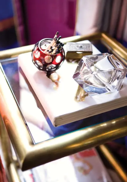 A faceted glass ashtray sits atop a closed book, next to a small silver box and a red glass match holder filled with matches.