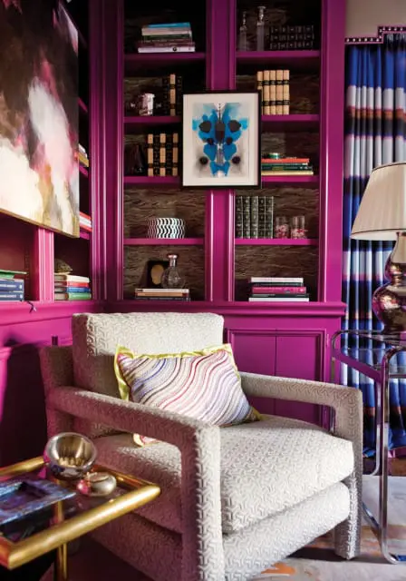Living room with magenta built-in bookshelves filled with books and decorative items, a patterned armchair with a striped pillow, and blue striped curtains.